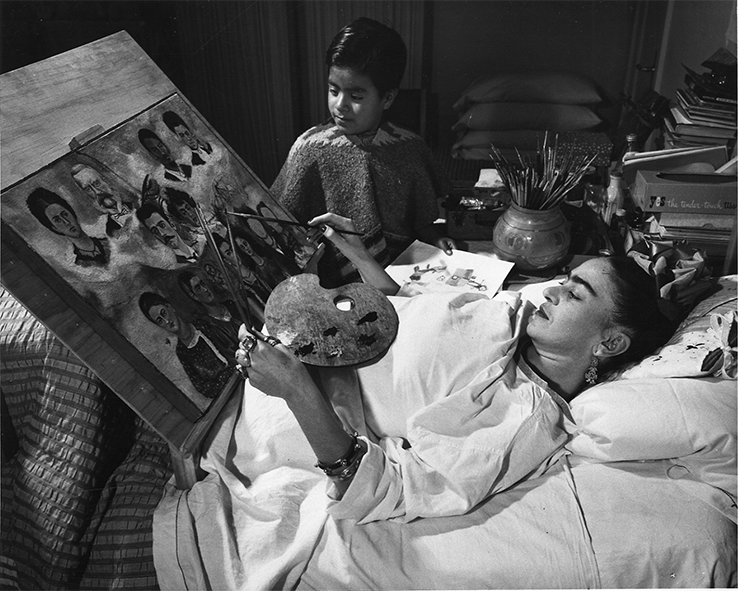 Une photo noir et blanc montre Frida Kahlo allongée dans son lit en train de peindre. Un jeune garçon la regarde