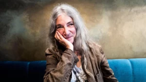 un portrait en couleur de Patti Smith souriant, assise sur un canapé bleu. Photo de Steve Peter